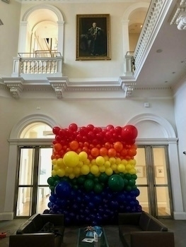 A large, colorful balloon display in a rainbow pattern is set up in an elegant room with archways and a portrait hanging above.