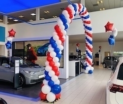 A car showroom is decorated with a large arch of red, white, and blue balloons.