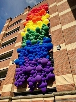 A vertical installation of colorful balloons arranged in a rainbow pattern is attached to the side of a brick building.
