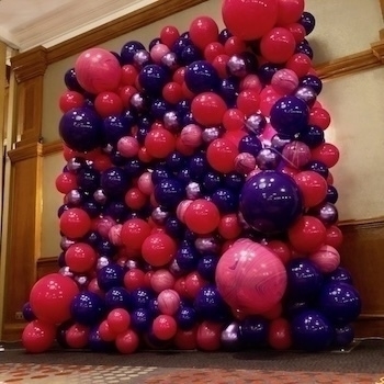 A wall of clustered balloons in shades of pink, purple, and blue is displayed in an indoor setting.