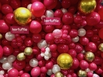 A colorful arrangement of red, pink, white, and gold balloons forms a festive backdrop with small kerfuffle signs.