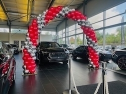 A showroom features a large balloon arch made of red and silver balloons, framing a row of cars.