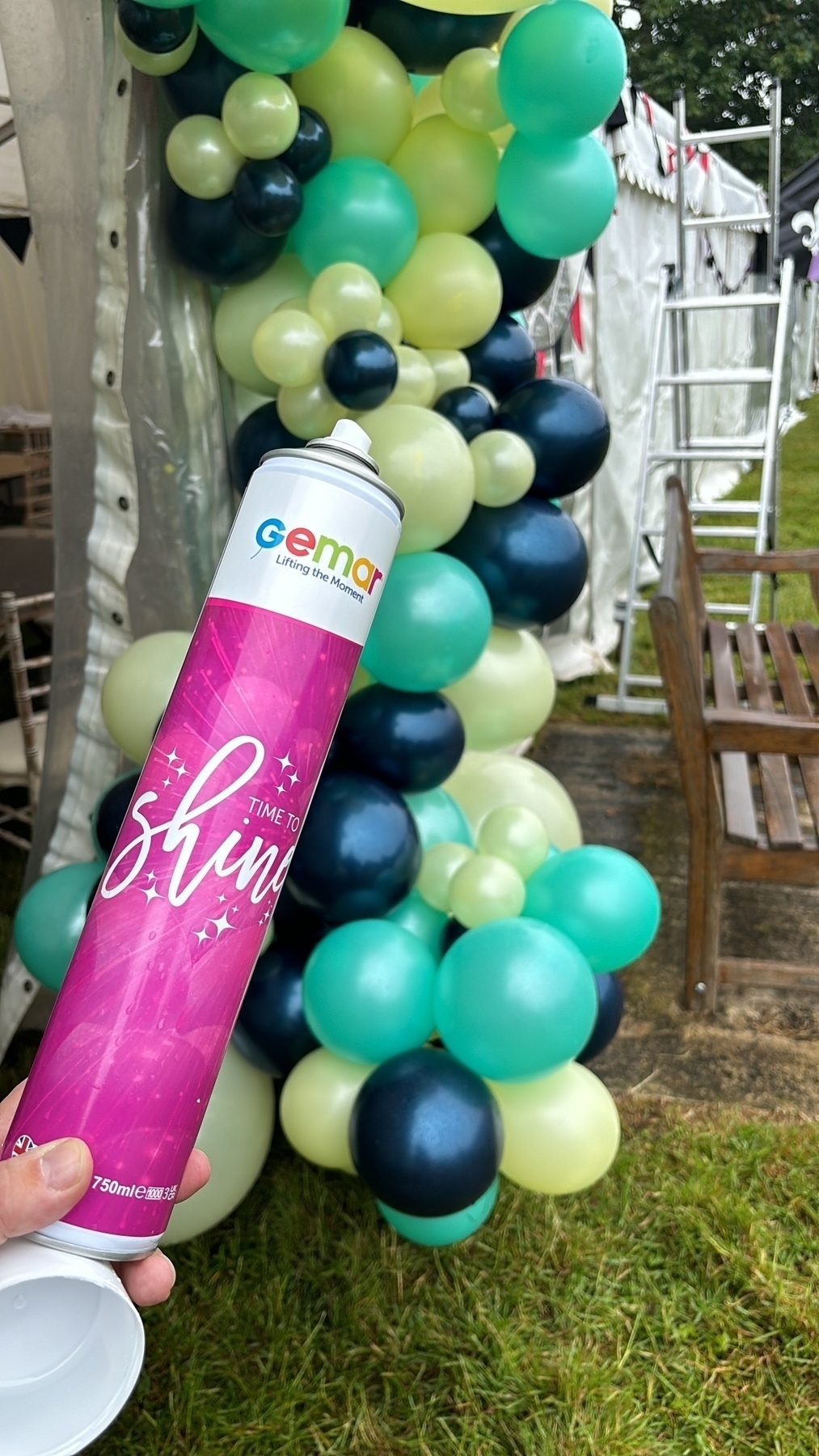 Auto-generated description: A hand holds a can labeled Time to Shine near a colorful balloon arrangement beside a tent and a wooden bench.