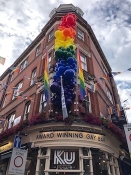 A building adorned with a large rainbow balloon decoration and flags has a sign indicating it's an award-winning gay bar.