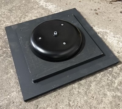 Auto-generated description: A black, circular object is mounted on a square base with a small protrusion in the center, placed on a rough, light-colored surface.